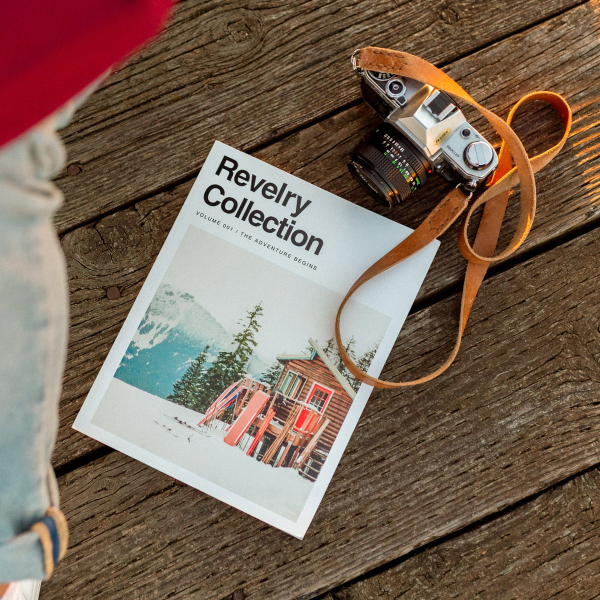 Person wearing a red coat and light blue jeans standing next to a magazine titled 'Revelry Collection' on a wooden surface.